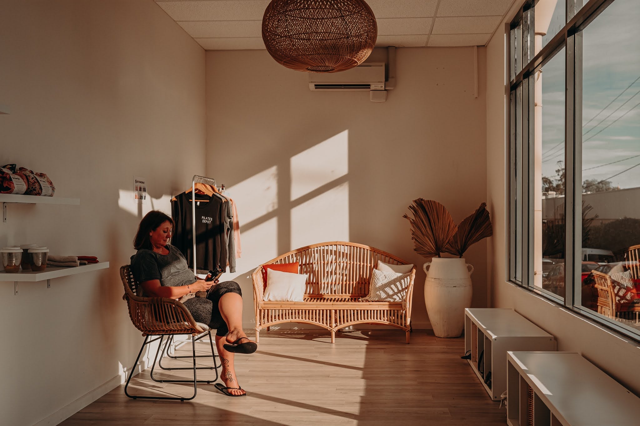 woman sitting in light filled room of the pilates depot studio in mount baker. waiting for a pilates reformer class. SHe is surrounded by TPD Store luxe pilates equipment on shelves