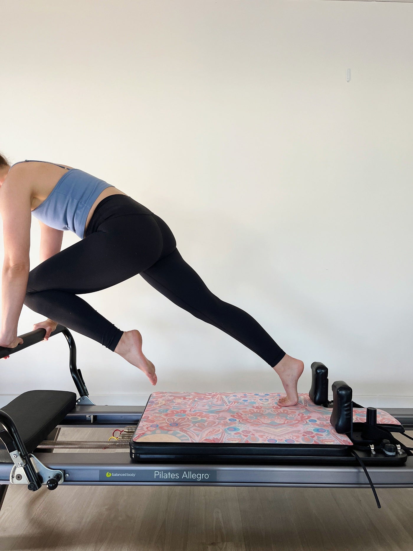 woman standing on pilates reformer machine that has a TPS Store eco friendly reformer mat in You are my sunshine Aurora Art collab print on the reformer bench. Woman is doing braced knee raises wuth hands on the bar and one foot on bench