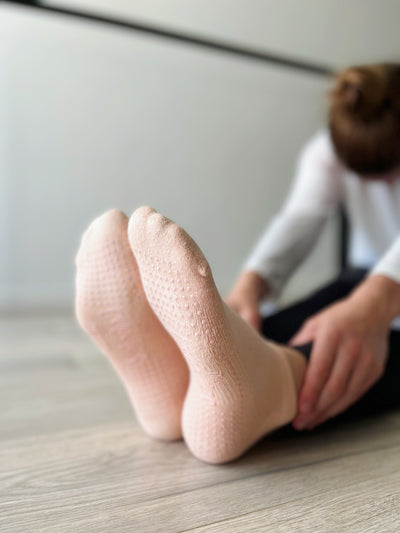 a pair of feet in peach fuzz low rise TPD store grippy socks. Woman leaning forward holding ankles.